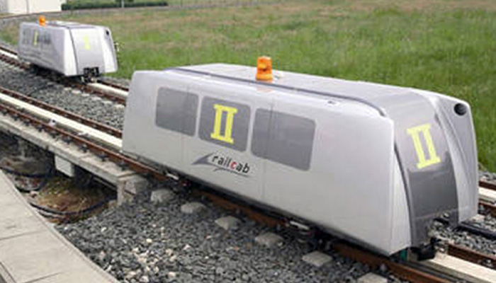Zwei kleine fahrerlose Züge mit der Aufschrift „railcab“ auf einem Gleis im Freien, umgeben von Gras und einem Kiesbett.