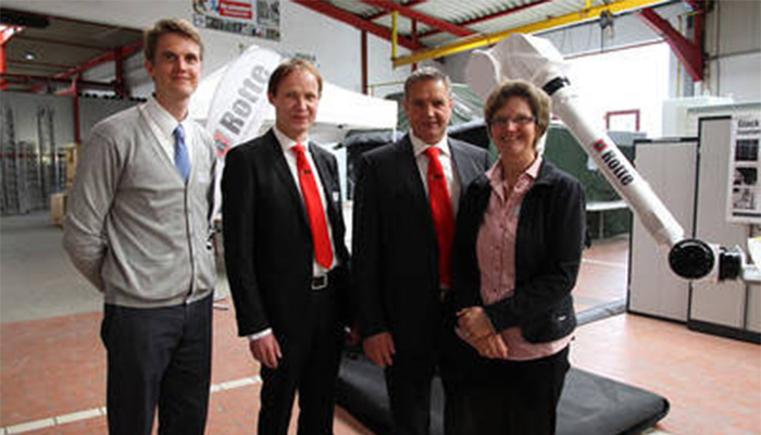 Vier Personen stehen nebeneinander in einer Halle vor einem großen weißen Industrieroboterarm mit der Beschriftung "Kuka".