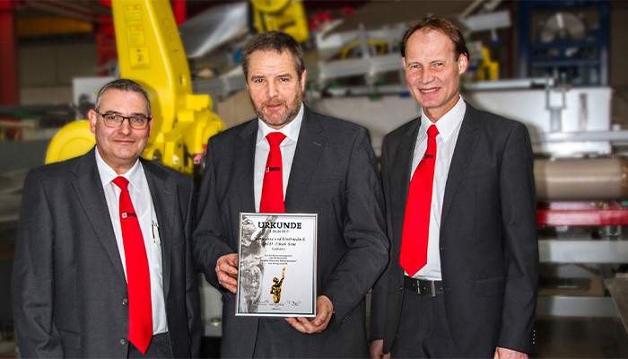 Drei Männer in Anzügen mit roten Krawatten stehen in einer industriellen Umgebung, einer hält eine Urkunde in der Hand.