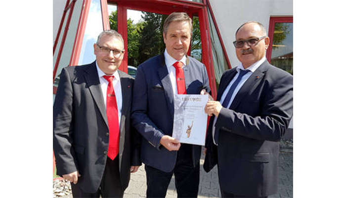 Drei Männer in Anzügen und roten Krawatten stehen vor einem Gebäude, der mittlere Mann hält eine Urkunde in der Hand.