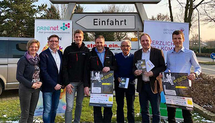 Sieben Menschen stehen vor einem Verkehrsschild mit der Aufschrift 'Einfahrt' und halten Informationsmaterial, im Hintergrund Werbebanner und ein parkendes Fahrzeug.