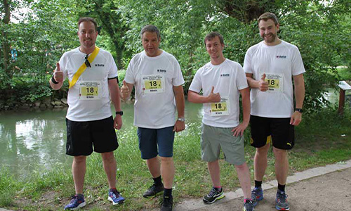 Vier Männer in weißen T-Shirts mit Laufnummern stehen nebeneinander auf einem Weg neben einem Gewässer und zeigen Daumen nach oben, umgeben von grünem Laub.
