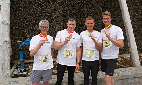 Vier Männer in weißen T-Shirts mit Startnummern und Medaillen um den Hals stehen vor einer hölzernen Struktur und halten ihre Medaillen in der Hand.