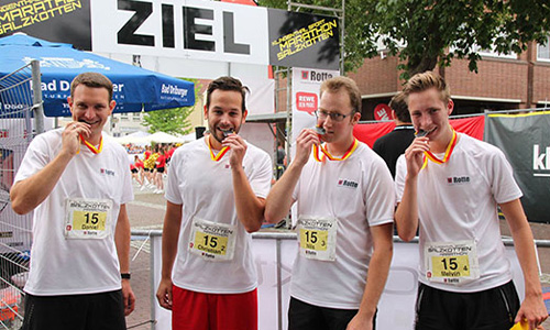 Vier Männer in weißen Laufshirts bei einer Ziellinie mit Medaillen im Mund, sie tragen Startnummern mit der Zahl 15 bei einem Laufwettbewerb.