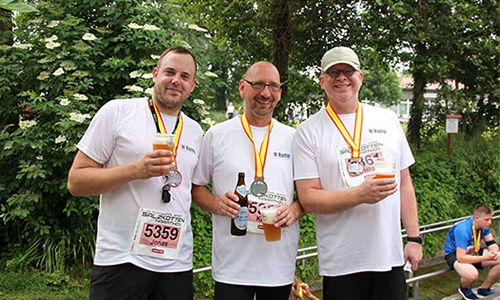 Drei Männer in weißen T-Shirts mit Laufnummern und Medaillen um den Hals stehen im Freien, jeder hält ein Getränk in der Hand, im Hintergrund Bäume und eine weitere Person auf einer Bank.