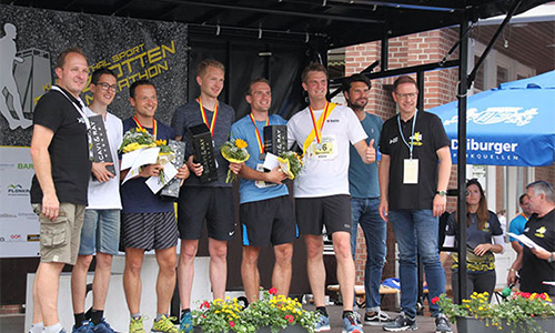 Sieben Männer stehen auf einer Bühne bei einer Sportveranstaltung, mehrere halten Blumen und Trophäen, hinter ihnen ein Banner mit der Aufschrift 
