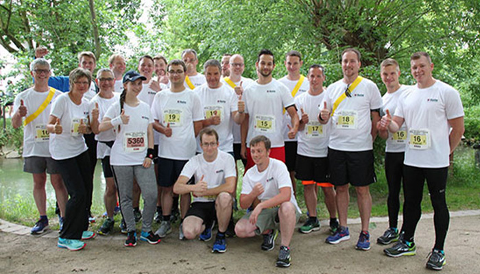 Gruppe von etwa 20 Personen in weißem Laufshirt mit Startnummern, die draußen vor grünen Bäumen und einem Fluss posieren und Daumen hoch zeigen.