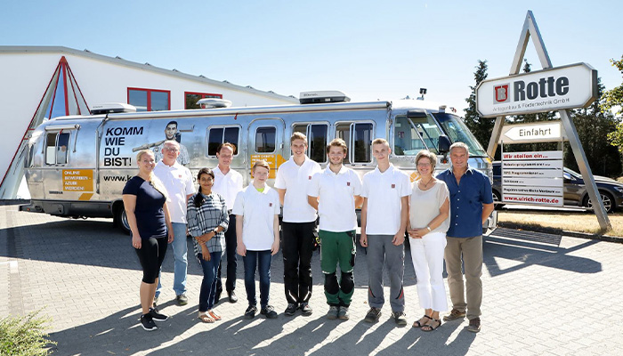 Gruppenfoto von neun Personen, darunter jüngere und ältere, stehend vor einem silbernen Anhänger mit Werbemotiv und neben einem Firmen-Schild mit der Aufschrift 'Rotte', draußen auf gepflastertem Gelände bei sonnigem Wetter.