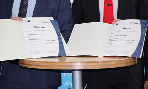 Zwei Personen in Anzügen halten auf einem Stehtisch zwei Urkunden mit blauen Rändern und unterschriebenen Feldern sichtbar nach vorne.