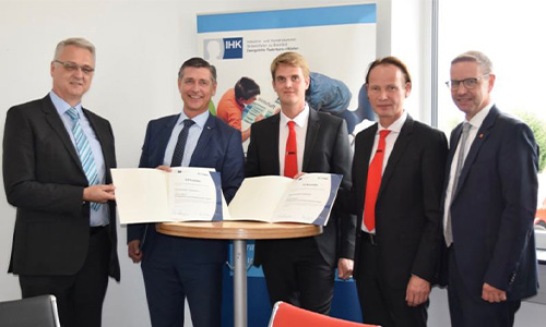 Fünf Männer in Anzügen, zwei davon halten offene Urkunden vor einem IHK-Banner in einem hellen Konferenzraum.