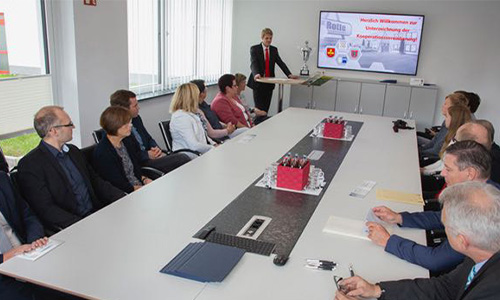 Konferenzraum mit Personen in formeller Kleidung, die an einem langen Tisch sitzen, während ein Mann am Kopfende einen Vortrag hält und Informationen auf einem Bildschirm zeigt.