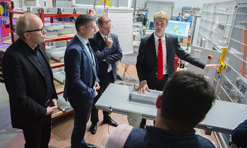 Fünf Männer in formeller Kleidung stehen um eine Maschine oder ein Gerät in einem industriellen Raum, wobei einer der jüngeren Männer mit roter Krawatte die Gruppe zu erklären scheint.