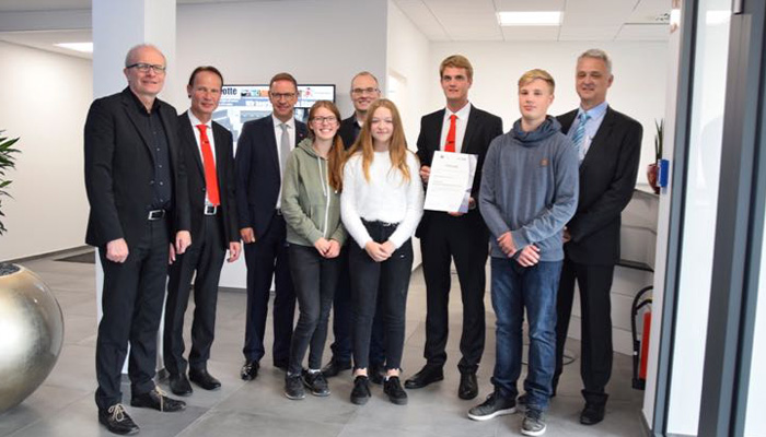 Gruppe von neun Personen, bestehend aus sechs Männern im Anzug oder Freizeitkleidung und drei Jugendlichen, eine hält ein Dokument, sie stehen in einem modernen Büroflur mit weiß gestrichenen Wänden und grauem Boden.