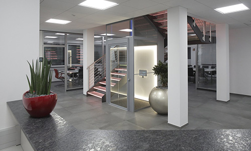 Moderner Büroflur mit Treppenhaus hinter einer Glasscheibe, zwei großen weißen Säulen, einem roten Pflanzkübel mit grüner Pflanze auf einer grauen Oberfläche und einem großen silbernen Pflanzgefäß neben den Treppen.
