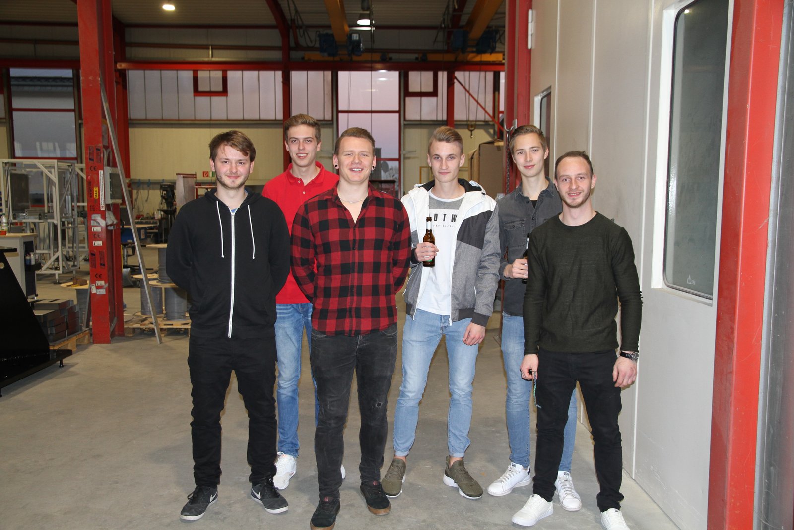 Sechs junge Männer stehen in einer Industriehalle vor einer roten Stahlkonstruktion, zwei von ihnen halten Bierflaschen, sie tragen legere Kleidung mit Jeans und Pullovern oder Hemden.