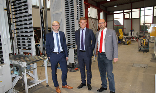Drei Männer in Anzügen stehen in einer Industriehalle mit Maschinen und Förderanlagen, im Hintergrund große Fenster und Produktionsausrüstung sichtbar.