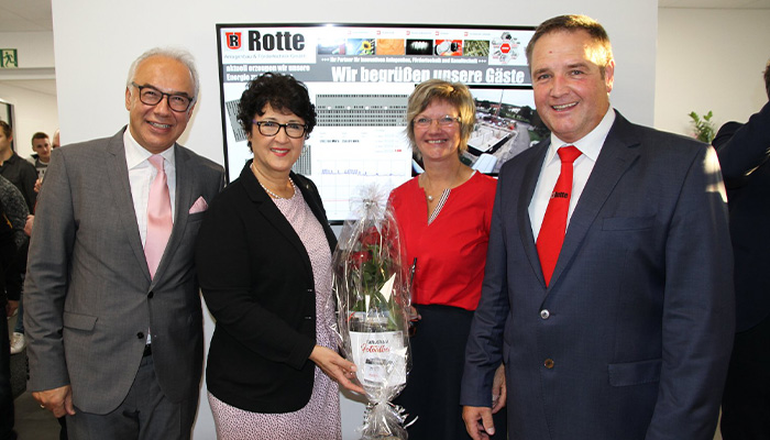 Vier lächelnde Personen in formeller Kleidung vor einem Bildschirm mit dem Text 'Rotte' und 'Wir begrüßen unsere Gäste', eine Frau hält einen verpackten Blumenstrauß