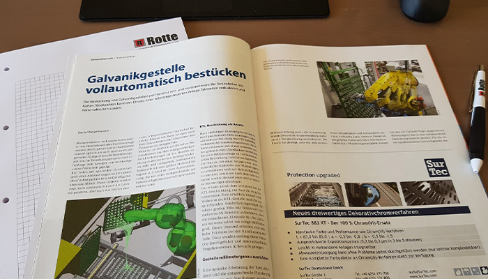 Zeitschriften- oder Zeitungsartikel auf einem Schreibtisch mit dem Titel "Galvanikgestelle vollautomatisch bestücken" in deutscher Sprache; der Artikel enthält Textspalten und mehrere Bilder von technischen Vorrichtungen, darunter grüne und gelbe mechanische Arme und industrielle Ausrüstung; daneben liegt ein Stift mit dem Aufdruck "SurTec".