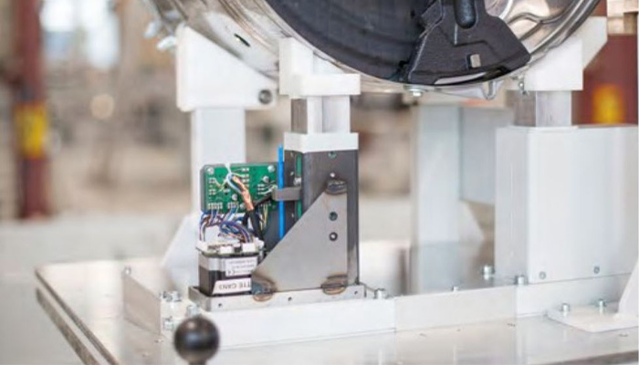 Nahaufnahme eines technischen Geräts mit Leiterplatte, Drähten und Metalhalterung auf weißem Untergrund in einer Werkstattumgebung.