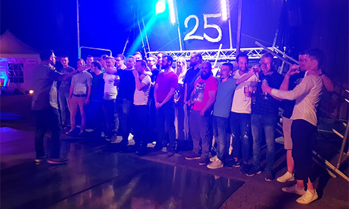 Gruppe von etwa 20 Personen, hauptsächlich Männer, stehen nebeneinander auf einer Bühne oder vor einem Podest mit einer Beleuchtung, die eine große 