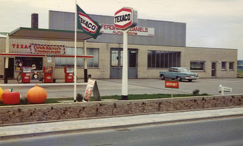 Außenansicht einer alten Texaco-Tankstelle mit zwei Texaco-Schildern, einer klassischen Zapfsäule, einem blauen Oldtimer-Auto auf dem Hof und Kürbissen vor dem Gebäude an einem bewölkten Tag.
