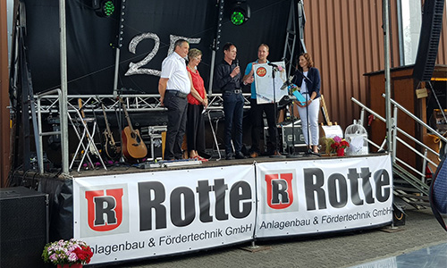 Fünf Personen stehen auf einer Bühnenplattform mit einem Banner der Firma Rotte Anlagenbau & Fördertechnik GmbH, eine Person hält ein Trikot mit der Nummer 25, im Hintergrund sind eine Gitarre und andere Bühnentechnik zu sehen.