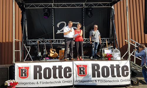 Drei Personen stehen auf einer Bühne mit Gitarren und Technik im Hintergrund, zwei Banner mit der Aufschrift 