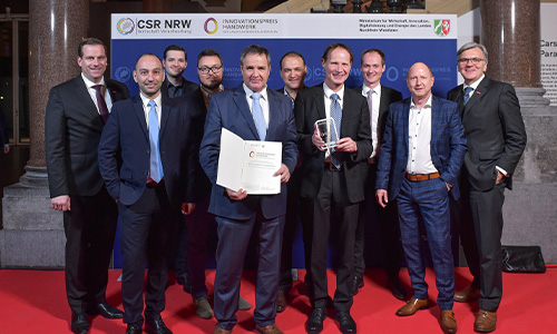 Neun Männer in Anzügen stehen auf einem roten Teppich vor einer Fotowand mit Logos von CSR NRW und Innovationspreis Handwerk, zwei Männer in der Mitte halten eine Urkunde und eine Trophäe.