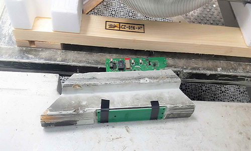 Eine abgebaute Aluminiumhalterung liegt auf einem weißen Tisch, dahinter sind elektronische Bauteile mit grünen Platinen und einem schwarzen Chip sichtbar, im Hintergrund befindet sich ein Holzkasten mit der Aufschrift CZ-056-HT und eine graue Schlauchleitung.