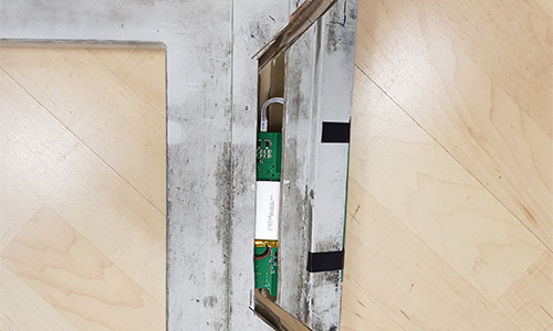 Teilweise geöffnete Metallabdeckung auf einem Holzfußboden, darunter sichtbar eine grüne Leiterplatte mit elektronischen Komponenten und einem weißen Modul.
