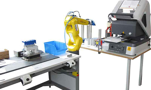 Industrieroboterarm in Gelb, der zwischen einem Förderband und einer Prüfmaschine auf einem Tisch positioniert ist, vor weißem Hintergrund