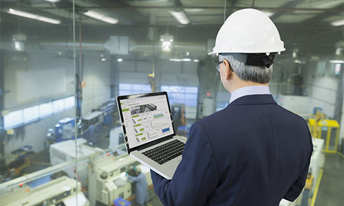 Person mit weißem Schutzhelm und Anzug, die einen Laptop in einer Industriehalle oder Fabrikumgebung betrachtet