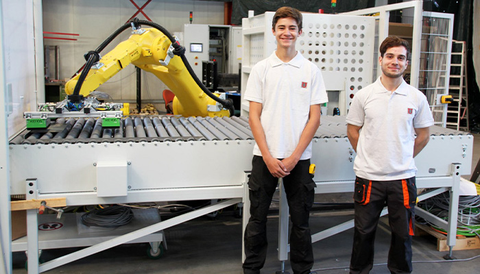 Zwei junge Männer in weißen Poloshirts und schwarzer Arbeitskleidung stehen vor einer industriellen Förderanlage mit einem gelben Industrieroboterarm in einer Werkstatt oder Fabrikumgebung.
