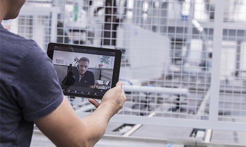 Person in T-Shirt hält ein Tablet mit einem Videoanruf eines Mannes, im Hintergrund eine industrielle Umgebung mit Metallgittern und Maschinen