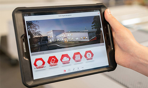 Hand hält ein Samsung-Tablet mit einer Bildschirmansicht einer digitalen Arbeitsplattform, die eine Gebäudeabbildung und verschiedene rot-weiße Icons zeigt