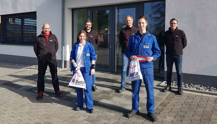Sechs Personen vor einem Gebäude, zwei vorne in blauen Arbeitsanzügen mit Tüten, vier hinten in schwarzer und dunkler Kleidung, alle schauen in die Kamera.