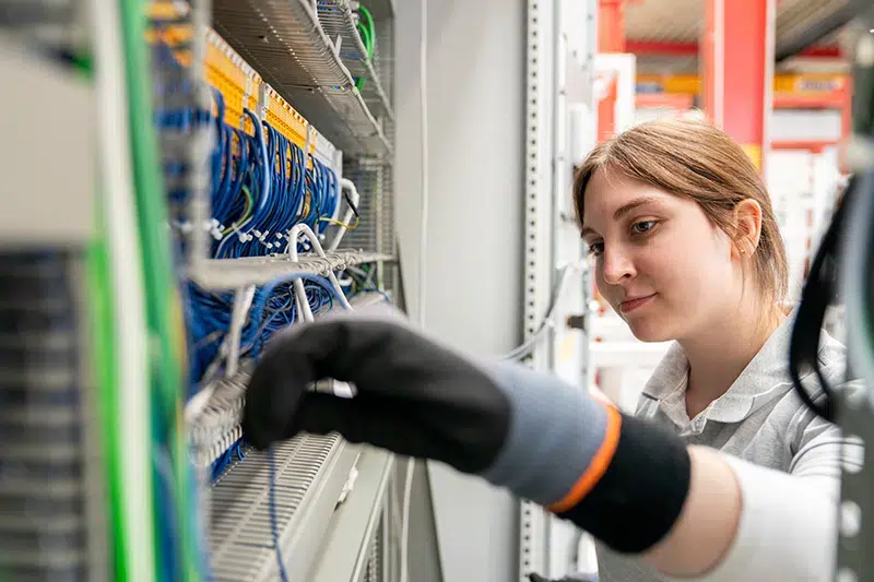 Junge Frau mit grauem Hemd und schwarzer Schutzhandschuh arbeitet konzentriert an einem Netzwerk-Schaltschrank mit vielen blauen Kabeln in einem Rechenzentrum oder Serverraum.