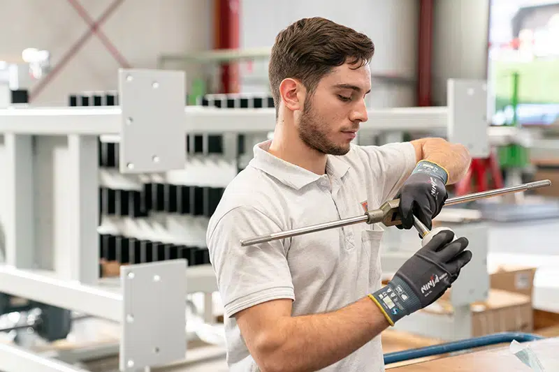 Junger Mann mit Sicherheits-Handschuhen, der in einer Werkstatt eine Stange aus Metall hält und inspiziert, umgeben von Werkstatt-Equipment im Hintergrund.