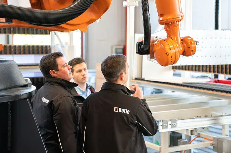Drei Männer in schwarzen Jacken mit Firmenlogo stehen in einer Industriehalle vor einem orangen Industrieroboter und beobachten diesen sorgfältig.