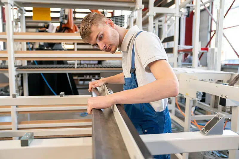 Ein junger Mann in blauer Arbeitslatzhose und weißem T-Shirt arbeitet mit einer Aluminiumstange an einer Maschine in einer Werkstatt.