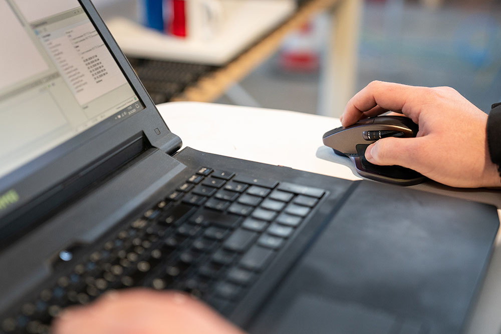 Nahaufnahme einer Person, die an einem Laptop arbeitet, mit einer Hand auf einer kabellosen Computermaus auf einem weißen Tisch.