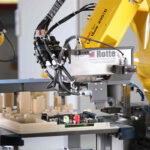 Gelbe Industrieroboterarme mit verschiedenen Kabeln und Präzisionswerkzeugen, die elektronische Bauteile und Prototypen auf einer Werkbank in einem Labor oder Fertigungsbereich bearbeiten.