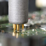 Nahaufnahme einer automatisierten Prüf- oder Lötspitze mit mehreren goldfarbenen Nadeln, die auf einer grünen Leiterplatte positioniert ist, um elektronische Bauteile zu testen oder zu verbinden.