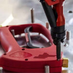 Nahaufnahme eines roten Handgriffes, an dem eine automatisierte Maschine mit einem schwarzen Werkzeugaufsatz arbeitet, um eine Schraube zu befestigen, mit metallischen Zylindern auf einer Werkbank im Hintergrund
