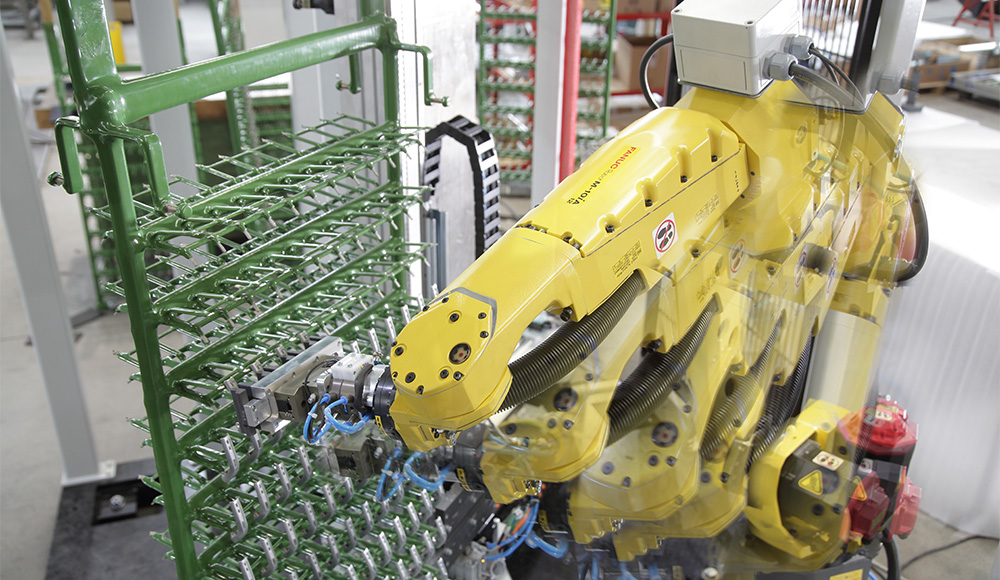 Gelber Industrieroboterarm, der an einem grünen Metallgestell mit vielen kleinen Halterungen arbeitet, mit Bewegungsunschärfe auf dem Arm, in einer hellen Fabrikumgebung.