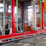 Produktionshalle mit einer großen, roten Industriemaschine, einem Mann in Arbeitskleidung und einer Metallleiter im Vordergrund.