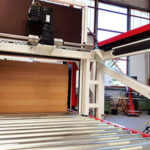 Industrieanlage mit einem automatisierten Fördersystem aus Metallrollen, das ein flaches, rechteckiges Holzpaneel in eine Maschine befördert, umgeben von einer hellen Werkhalle mit großen Fenstern.