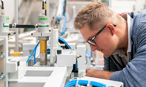 Junger Mann mit Brille inspiziert konzentriert eine komplexe Maschine oder Anlage in einem technischen Umfeld mit Kabeln und Metallteilen