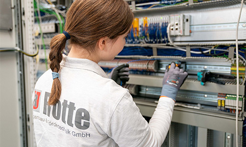 Person mit Zopf und Handschuhen arbeitet an einem offenen Schaltschrank mit vielen Kabeln und elektronischen Komponenten