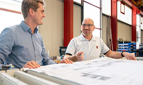 Zwei Männer, einer im blauen Hemd und einer im weißen Polohemd mit Brille, die über einen großen technischen Plan an einem Tisch in einer Industriehalle diskutieren